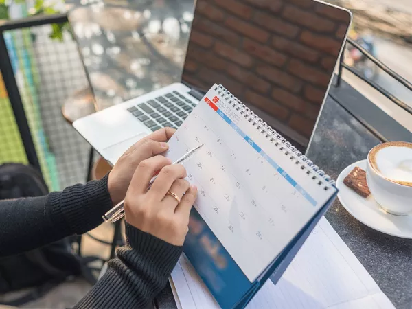 Hands holding a pen over a calendar, computer and coffee cup.