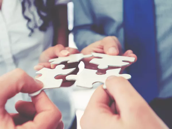 Four hands each holding a puzzle piece in a four corner square, about to put them together.