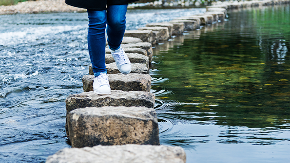 Pair of legs in blue jeans with white sneakers walking through steppingstones in a body of water