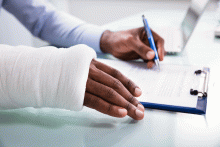 Overhead view of injured man with bandaged hand filling insurance claim form on clipboard.