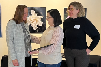 Two women from the Department of Children and Families receive the award certificate from Jen Flogel of the Department of Administration. All three women are standing, with photos of flowers behind them.